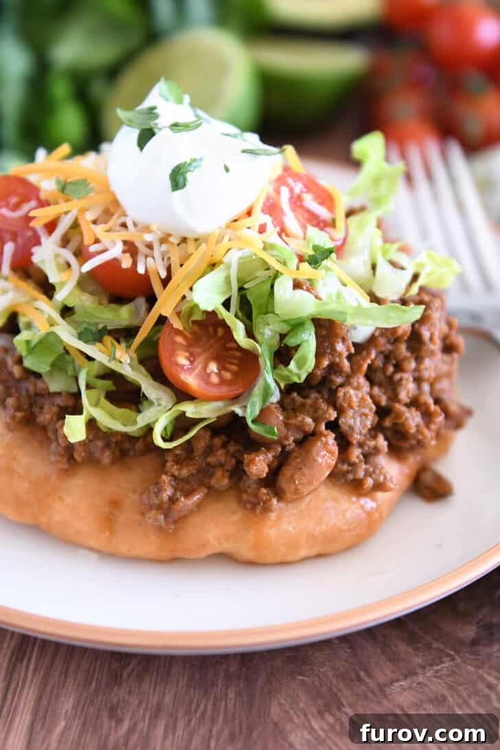 Piece of fry bread on cream plate topped with taco meat, lettuce, tomatoes, cheese, and sour cream.