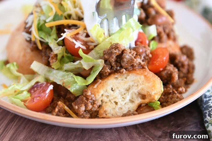 Piece of fry bread on cream plate topped with taco meat, lettuce, tomatoes, cheese, and sour cream with piece cut off on fork.