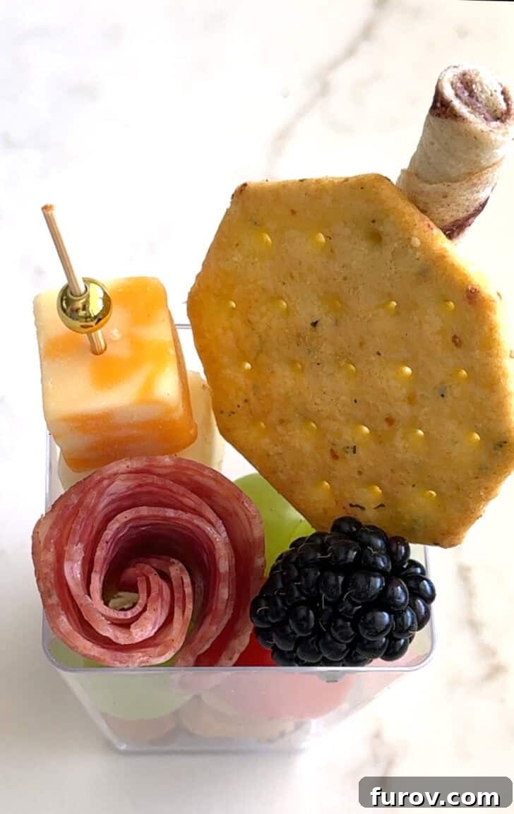 Square plastic cup with cracker, cheese skewer, rolled salami, blackberry, and pirouline.