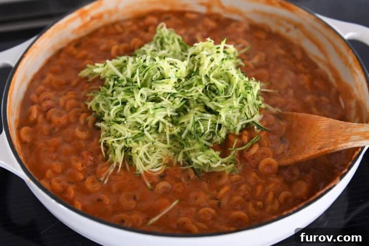 Adding shredded zucchini to cooked pasta in red sauce with shredded zucchini in deep skillet.